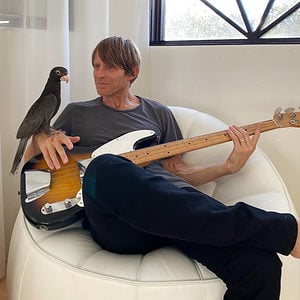 Chris Chaney sat in a chair with a bass guitar and a parrot on one hand.