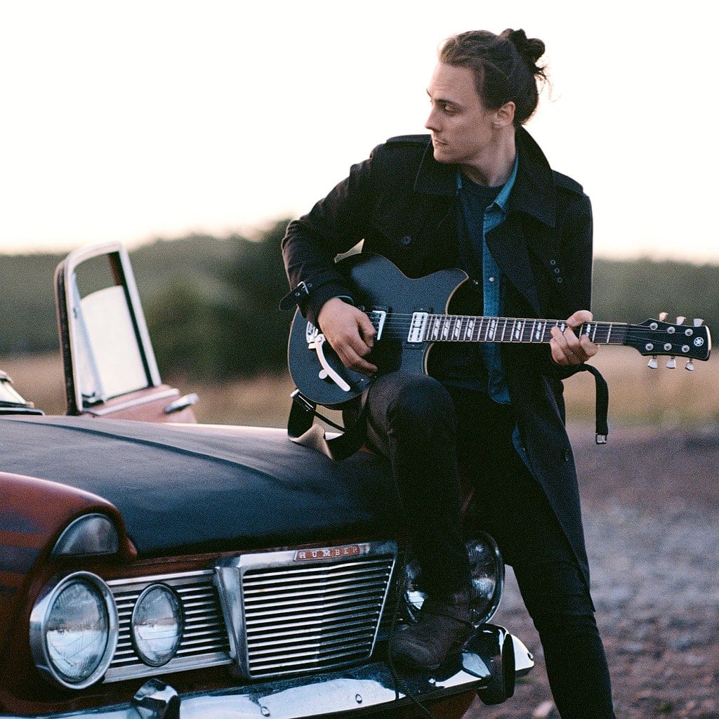 Chris Buck sat on a car bonnet playing guitar.