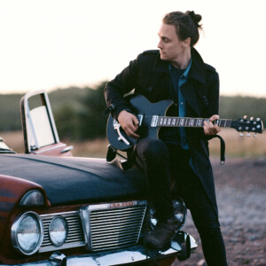 Chris Buck sat on a car bonnet playing guitar.